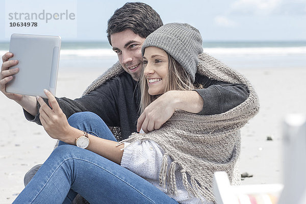 Ein Paar sitzt am Strand und nimmt einen Selfie mit digitalem Tablett.