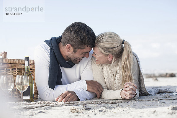 Lachendes Paar  das auf einer Decke am Strand liegt und Picknick macht.