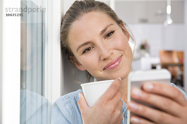 Junge Frau sitzt am Fenster  trinkt Kaffee und liest SMS