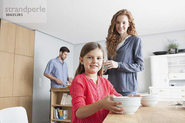 Porträt eines kleinen Mädchens  das den Tisch in der Küche mit seinen Eltern im Hintergrund deckt.