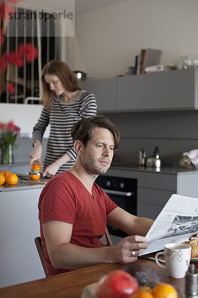 Mann liest Zeitung in der Küche  Frau macht Orangensaft im Hintergrund