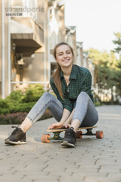 Porträt einer glücklichen jungen Frau  die auf einem Skateboard im Freien sitzt.