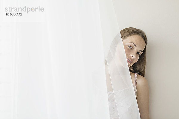 Porträt einer lächelnden Frau  die sich zu Hause hinter dem Vorhang versteckt.