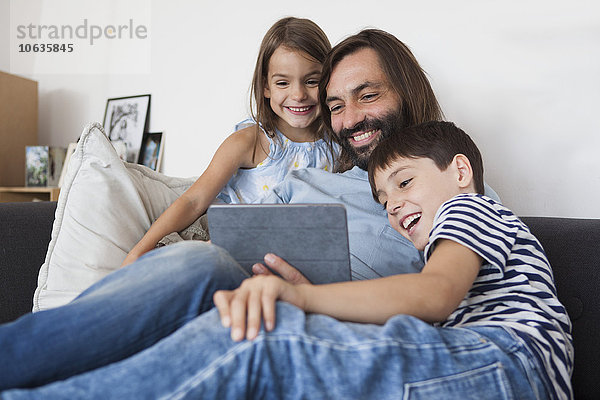 Glücklicher Vater mit Kindern  die zu Hause ein digitales Tablett auf dem Sofa benutzen.