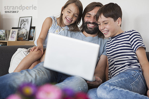 Fröhlicher Vater mit Kindern  die den Laptop zu Hause auf dem Sofa benutzen.