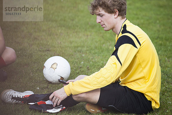 Junger Fußballspieler mit Handy im Sitzen auf dem Spielfeld