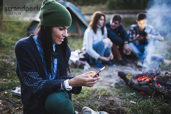 Junge Frau mit Handy  während Freunde am Lagerfeuer auf dem Campingplatz sitzen