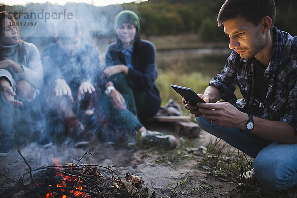 Junger Mann beim Sitzen mit Freunden am Lagerfeuer mit digitalem Tablett