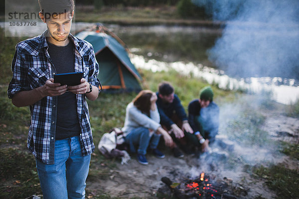 Junger Mann mit digitalem Tablett  während Freunde am Lagerfeuer im Wald sitzen.