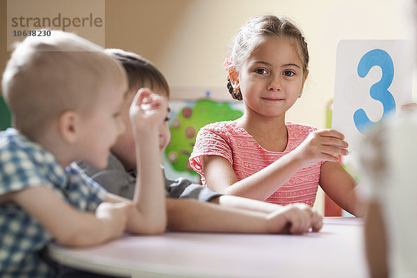 Porträt des süßen Mädchens mit Nummer 3 Plakat  das neben Freunden im Klassenzimmer sitzt