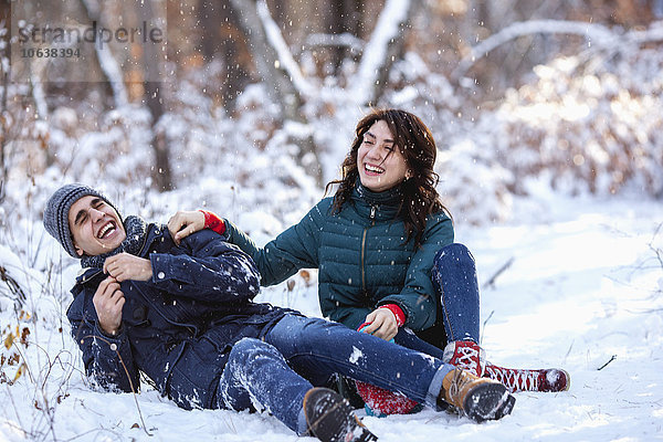Verspieltes junges Paar in voller Länge auf verschneitem Feld sitzend