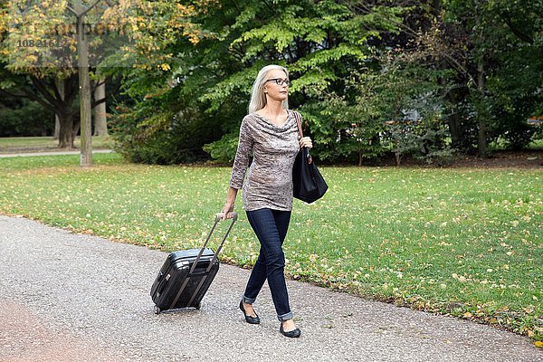 Reife Frau  die durch den Park läuft und den Koffer auf Rädern zieht.