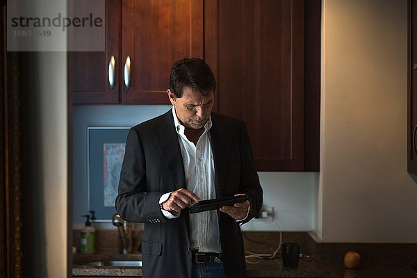 Älterer Mann in Anzugjacke in der Küche  hält digitales Tablett mit Blick nach unten.