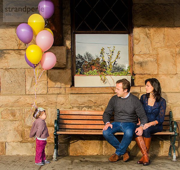 Junges Mädchen steht mit einem Haufen Luftballons  reifes Paar sitzt auf der Bank und beobachtet sie.