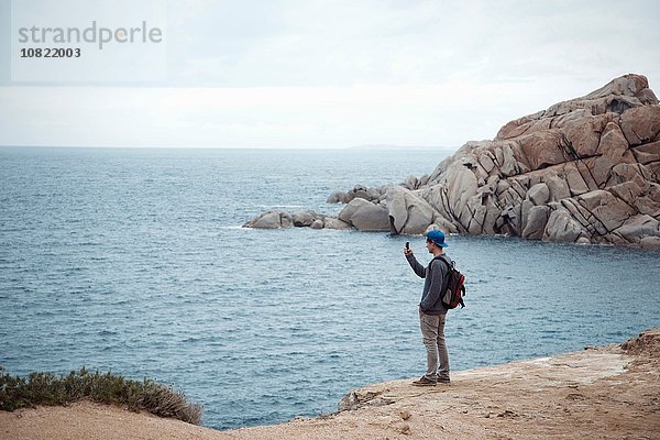 Junger Mann steht auf einer Klippe und fotografiert mit dem Smartphone den Ozean  Costa Smeralda  Sardinien  Italien