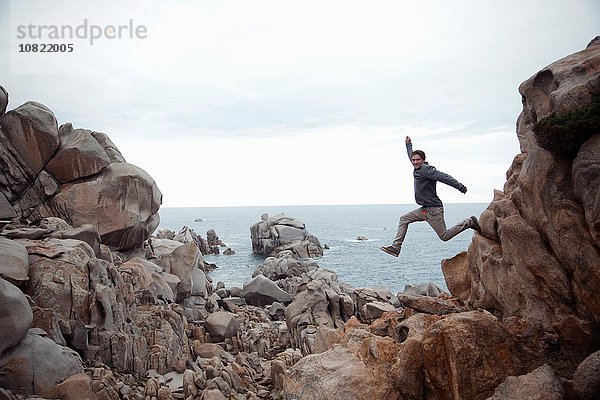 Seitenansicht eines jungen Mannes in der Luft  der von Felsen springt  lächelnd  Costa Smeralda  Sardinien  Italien