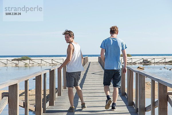 Durchgehende Rückansicht von jungen Männern  die auf einem hölzernen Pier laufen und über die Schulter schauen  Sardinien  Italien