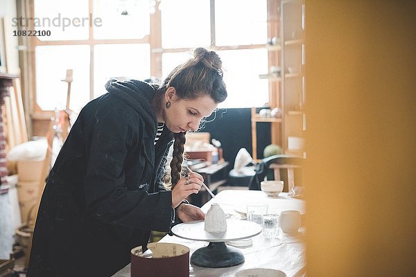 Seitenansicht der jungen Frau in der Werkstatt beim Auftragen der Keramikglasur auf den Tontopf  Blick nach unten