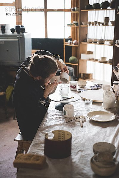 Seitenansicht eines erwachsenen Mannes  der in der Werkstatt sitzt und Keramikglasur auf den Tontopf aufträgt.