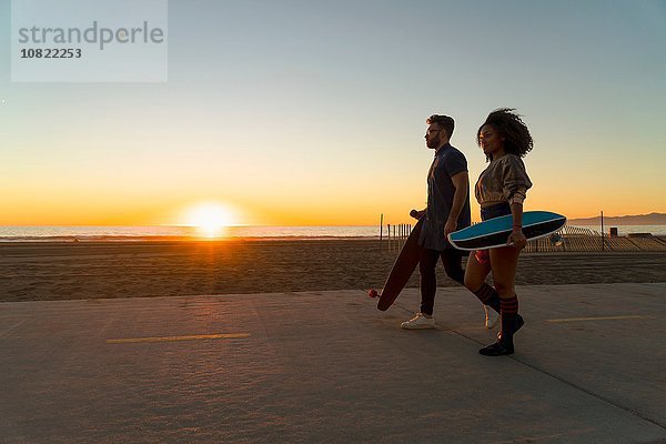 Paar  das am Strand entlang geht und Skateboards hält.