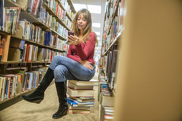 Reife Frau sitzt auf einem Bücherstapel in der Bibliothek und liest Smartphone-Texte.