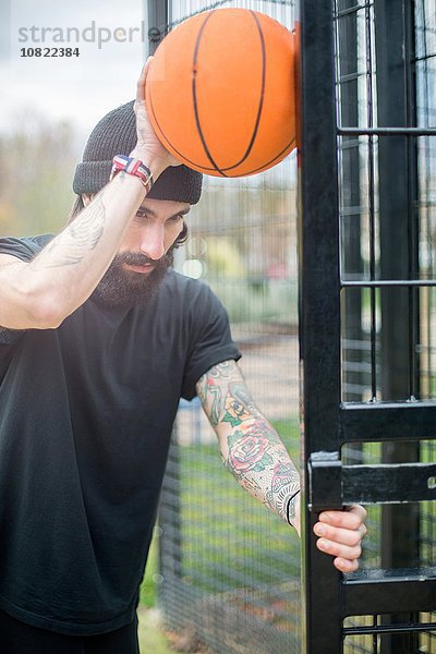 Mittlerer erwachsener Mann steht am Zaun  hält Basketball gegen Zaun  nachdenklicher Ausdruck