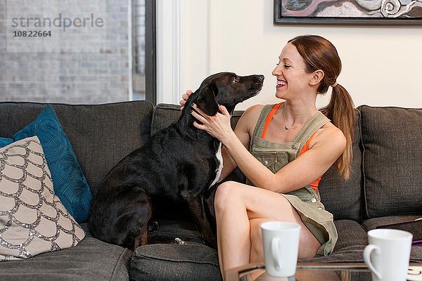 Junge Frau sitzt auf dem Sofa von Angesicht zu Angesicht mit lächelndem Hund.