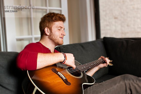 Junger Mann sitzt auf dem Sofa und spielt Gitarre  die Augen geschlossen.