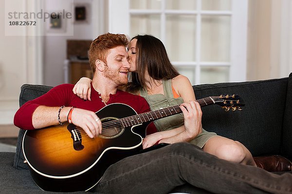 Junge Frau auf dem Sofa küsst jungen Mann  der Gitarre auf der Wange spielt.