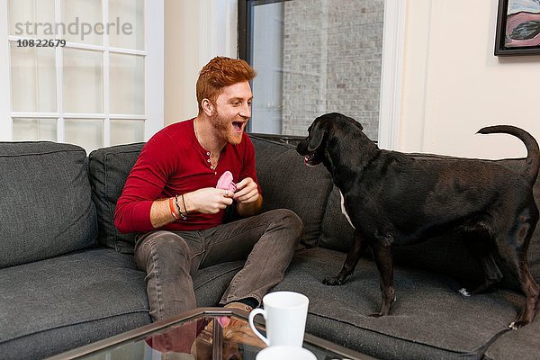 Junger Mann sitzt auf dem Sofa und spielt mit einem Hund  der den Mund aufmacht und lächelt.
