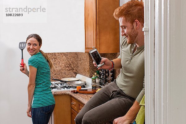 Junger Mann in der Küche mit dem Smartphone  um die junge Frau mit dem Spachtel zu fotografieren.