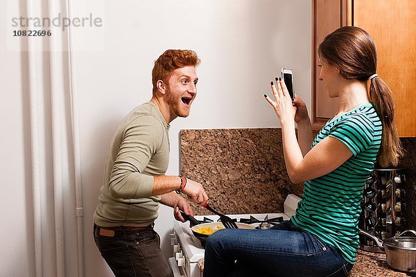 Seitenansicht der jungen Frau in der Küche mit dem Smartphone  um den jungen Mann beim Kochen zu fotografieren.