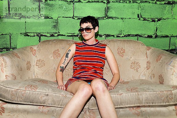 Porträt einer erwachsenen Frau mit Sonnenbrille  die auf einem getragenen Sofa sitzt.