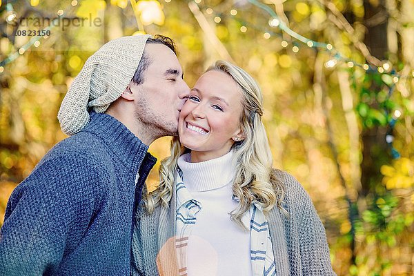 Porträt eines jungen Mannes  der eine junge Frau auf der Wange im Wald küsst.