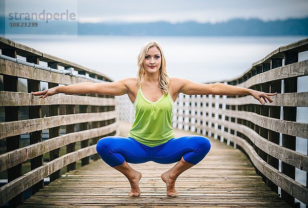 Porträt einer jungen Frau  die in Yoga-Pose auf einem hölzernen Pier hockt.