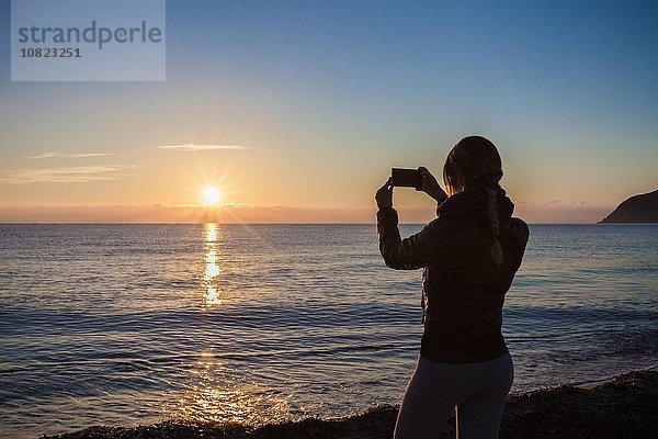 Hintere Silhouette einer jungen Frau  die den Sonnenuntergang über dem Meer fotografiert  Villasimius  Sardinien  Italien