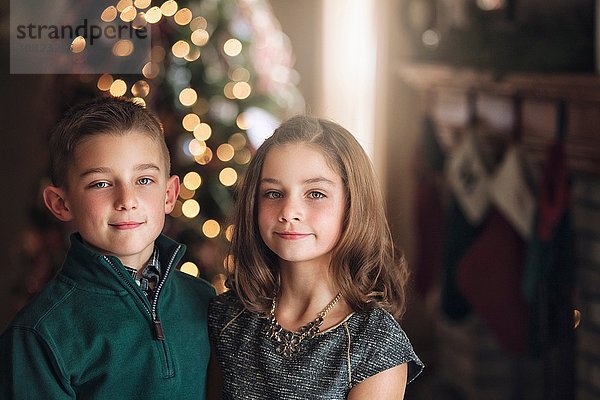 Porträt von Mädchen und Junge vor dem Weihnachtsbaum mit lächelndem Blick auf die Kamera