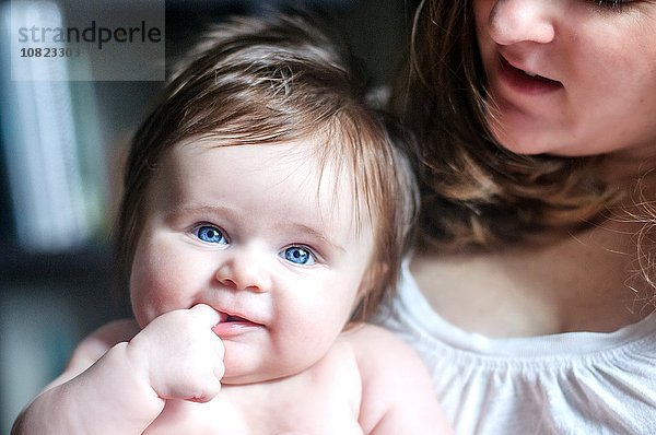 Mutter hält Baby Tochter mit Finger im Mund und schaut lächelnd in die Kamera.