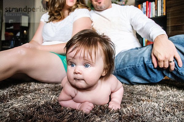 Mutter und Vater sitzen Rücken an Rücken auf einem Teppich  das Mädchen liegt auf einem Teppich und schaut weg.