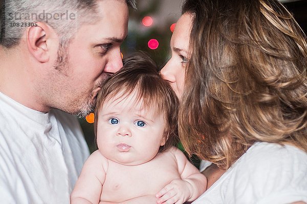 Mutter und Vater von Angesicht zu Angesicht haltendes kleines Mädchen  das sich auf den Kopf küsst.