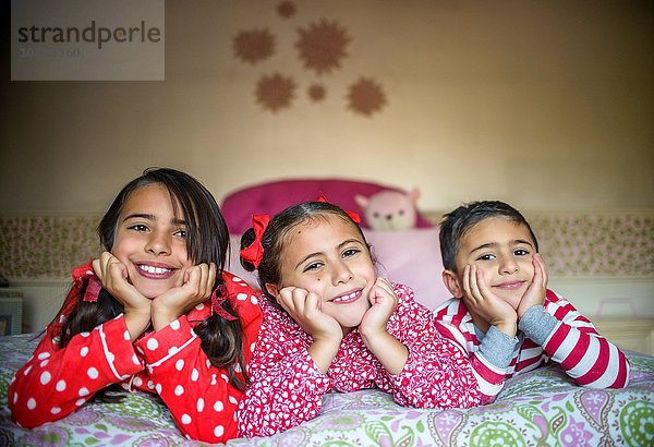 Kinder im Schlafanzug liegend auf dem Bett Kinn in den Händen und schauen lächelnd in die Kamera.