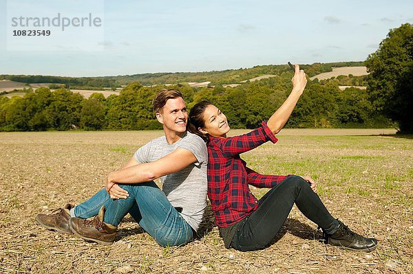 Junges Wanderpaar sitzt Rücken an Rücken im Feld und nimmt Smartphone Selfie  Great Missenden  Buckinghamshire  U.K.