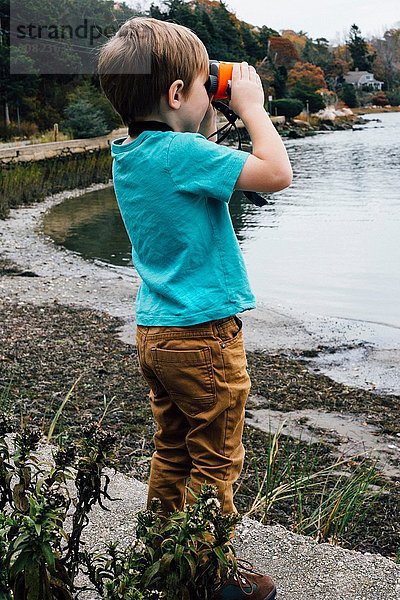 Kleiner Junge steht am See und schaut durch ein Fernglas.