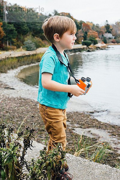 Kleiner Junge steht am See und hält ein Fernglas.