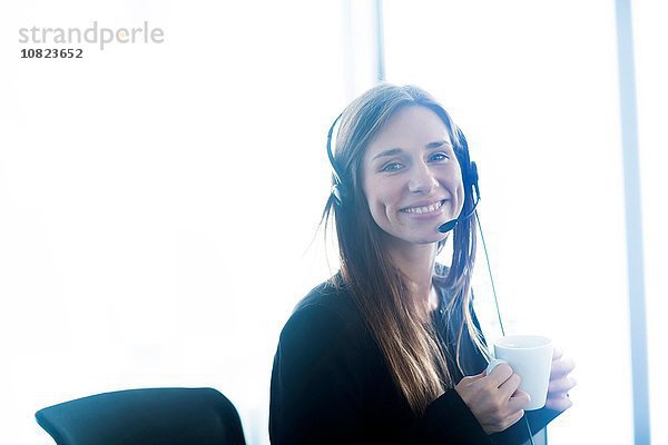 Junge Frau im Telefon-Headset mit Kaffeetasse und lächelndem Blick auf die Kamera