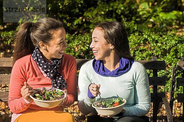 Junge erwachsene weibliche Zwillinge sitzen auf der Parkbank und essen zu Mittag.