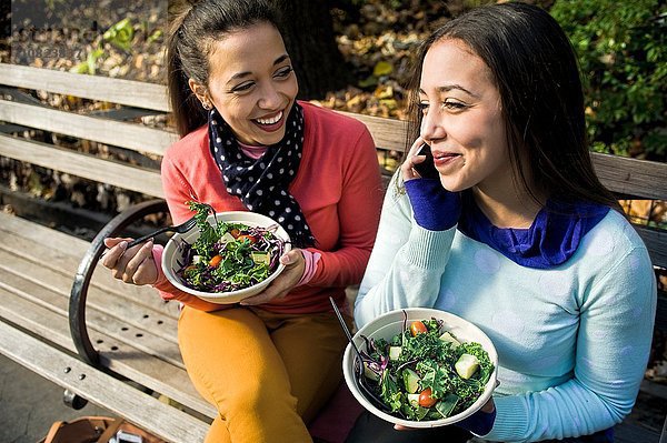 Junge erwachsene weibliche Zwillinge sitzen auf der Parkbank  plaudern auf dem Smartphone und essen zu Mittag.