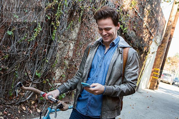 Junger Mann schlendert mit dem Fahrrad auf dem Bürgersteig und liest Smartphone-Texte.