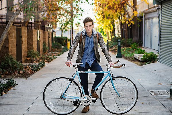 Porträt eines jungen Mannes  der sich auf dem Bürgersteig gegen das Fahrrad lehnt.
