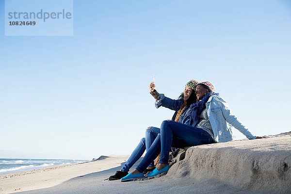 Zwei Freunde  die am Strand sitzen  sich selbst porträtieren  Smartphone benutzen